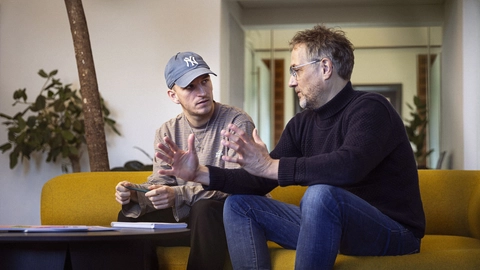 To personer, der sidder på en gul sofa, engageret i en samtale, mens de gestikulerer med hænderne, med planter i baggrunden og et bord med bøger og notater foran dem.